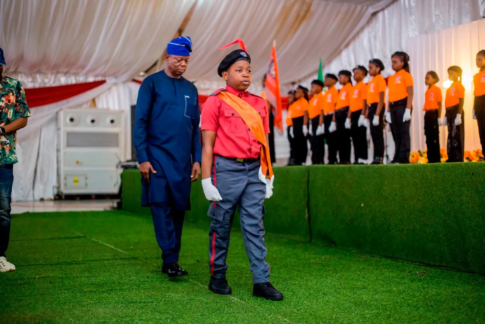A pupil in a red and grey cadet uniform with an orange sash marching beside a guest, past a line of pupils in orange