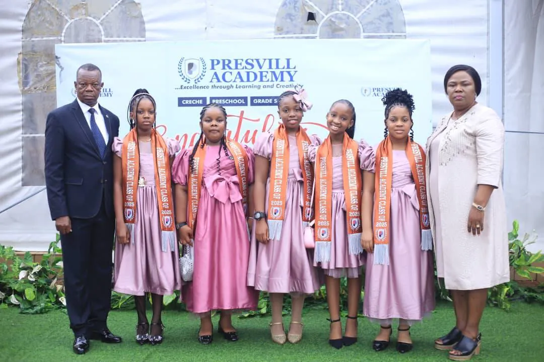 Graduating girls in pink dresses and orange sashes lined up in front of the Presvill Academy banner