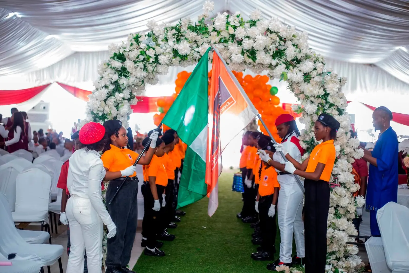 Pupils in orange and white carrying the Nigerian flag and the school flag beneath a floral arch