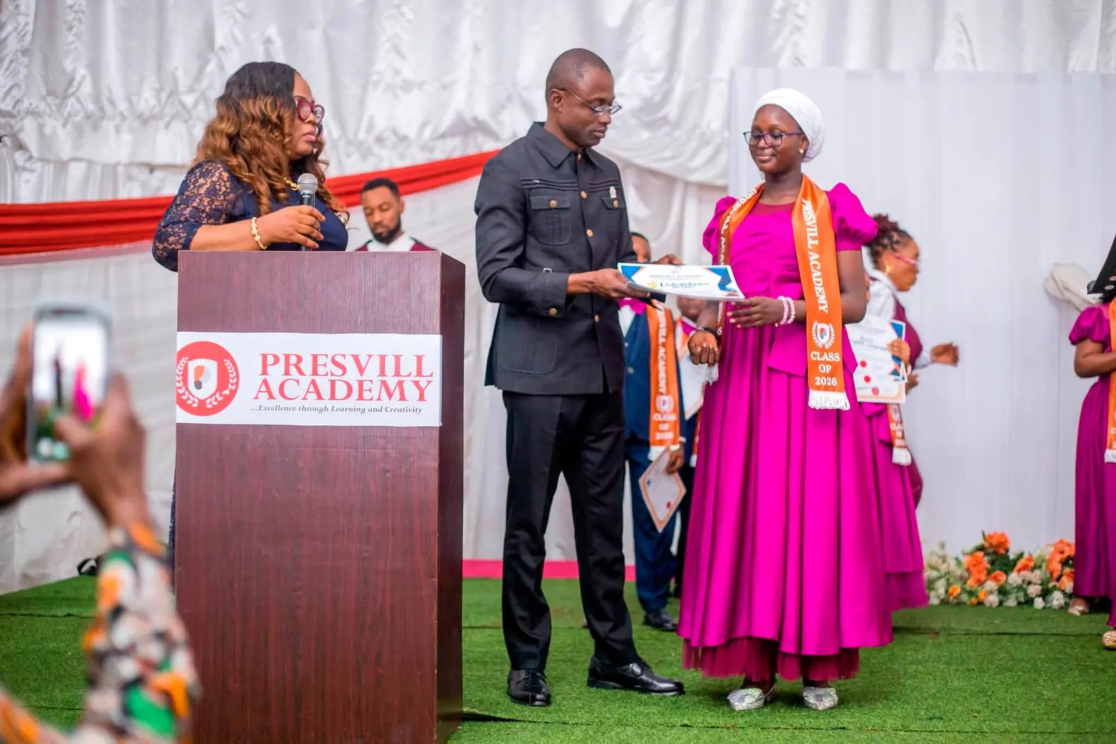 The 2026 valedictorian, in a pink gown and Class of 2026 sash, receiving a certificate beside the Presvill Academy lectern