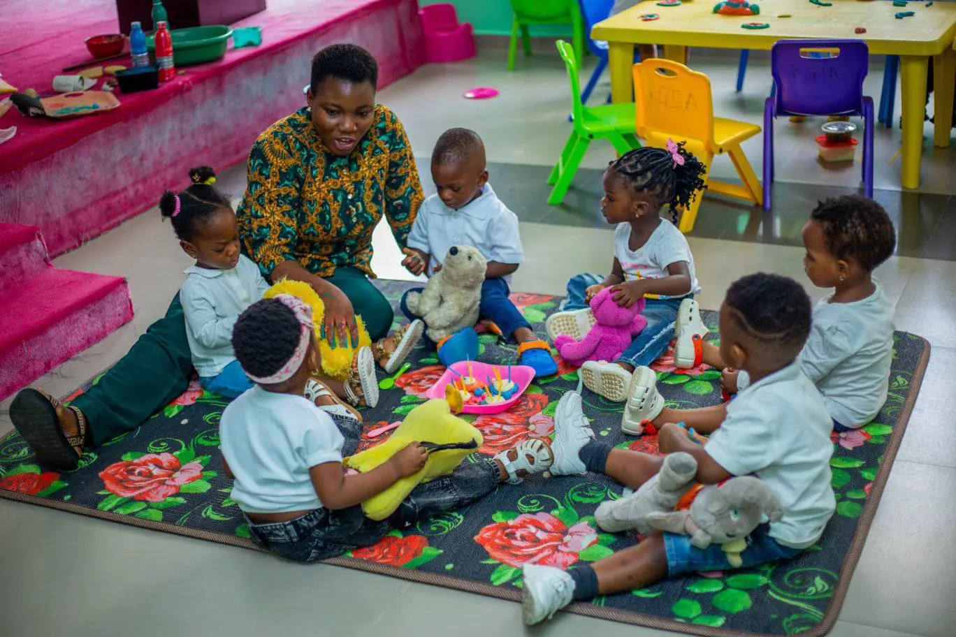 A Pre-School teacher sitting on a play mat with a small group of young children and their soft toys